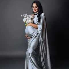 A full-length studio shot of an expectant mother in a graceful silver saree, holding a bouquet of white peonies. The long, flowing drape of the saree adds a touch of regal elegance to the portrait.
