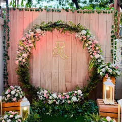 This rustic photo booth combines natural elements with personalized details. A large wreath of greenery and pastel flowers frames the couple's initials in a custom gold monogram. The wooden backdrop and lantern props complete the charming, rustic vibe.