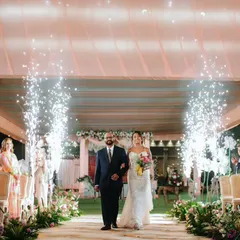 The bride, in a classic white wedding gown, is walked down the aisle by her father, with guests looking on and pyrotechnics lighting the way. A beautiful blend of traditions.