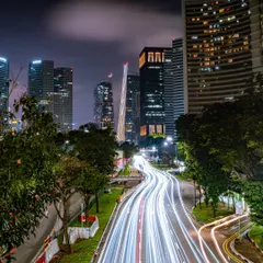 A long-exposure shot of a Singapore highway at night, transforming vehicle headlights and taillights into brilliant white streaks of light against the modern cityscape.