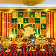 This traditional setup features a checkered backdrop in green and yellow, with hanging bells and marigold garlands. The striped floor mat adds to the classic, festive feel.