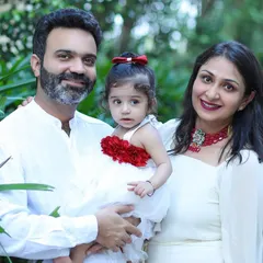 A classic family portrait taken outdoors, with a mother and father holding their daughter against a lush green background.