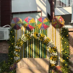 A beautiful outdoor Haldi setup featuring a floral arch of yellow and white marigolds against a backdrop of green and white striped drapes. The scene is brightened by colorful Rajasthani umbrellas suspended from above.