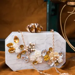 This white marble-effect acrylic clutch is adorned with golden-hued rhinestones and delicate 3D floral accents. It includes a detachable gold chain sling for versatile styling.