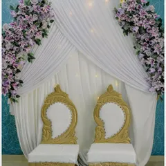 The elegant stage setup for an engagement ceremony. The white and gold chairs against a backdrop of white drapes and purple flowers create a regal look.