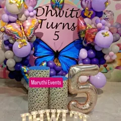 A stunning butterfly-themed decoration for Dhwiti's 5th birthday. The setup features a large central butterfly on the backdrop, surrounded by a purple and white balloon arch and large butterfly foil balloons.