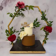 A sophisticated two-tier cake for a 25th silver jubilee anniversary, presented within a decorative golden hexagonal frame. The clean white top tier contrasts beautifully with the smooth brown bottom tier, adorned with fresh white roses.