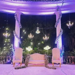 This elegant reception stage features white pillars wrapped in flowers, with crystal chandeliers hanging above the couple's seating. The surrounding glowing floral lights add a magical touch to the outdoor setting.
