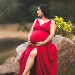 A stunning full-length portrait of a mother-to-be in a vibrant red dress, seated on a rock in a park. The natural green background makes the red color pop, creating a striking image.