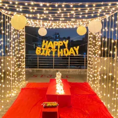 A romantic birthday dinner setup on a terrace. This decoration features a canopy of fairy lights, a low table covered in red fabric, and "Happy Birthday" foil balloons, creating a private and magical dining experience.