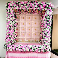 A beautiful pink and white floral frame designed for a home Ganpati setup. The backdrop is made of delicate white flowers with small golden bells, perfectly complementing the dense rose arrangement.