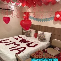 A personalized romantic setup with the name "UMA" written in rose petals on the bed. The room is filled with red and white ceiling balloons.