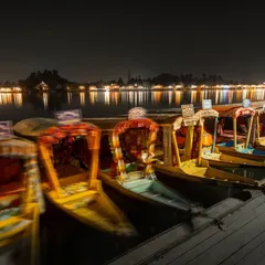 The shikaras and houseboats of Dal Lake in Srinagar, lit up at night. A long exposure captures the motion of the boats and the reflections on the water.