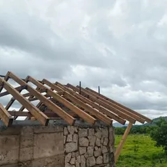 A building takes shape in harmony with its surroundings. Here, walls made of natural stone and rammed earth rise from the ground, while the wooden framework for the roof is put in place against a cloudy sky.