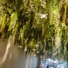 An enchanting ceiling installation for a lobby or entrance area. The dense hanging greenery and white flowers, interspersed with delicate glowing birds, creates a magical, forest-like atmosphere.