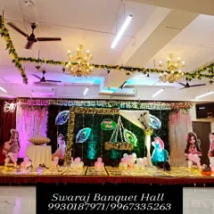 A wide view of a naming ceremony setup in our hall. The stage is decorated with a green turf backdrop, peacock feather elements, and a traditional cradle, with ample seating arranged for guests.