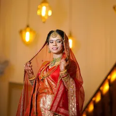 A portrait of a North Indian bride in a vibrant red saree, her veil delicately held. The warm indoor lighting creates a classic, timeless feel.