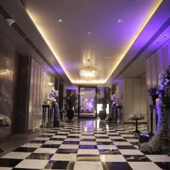 A chic and modern hotel corridor transformed into a grand entrance. The checkerboard floor, purple uplighting, and floral arrangements create a sophisticated and stylish pathway for guests.