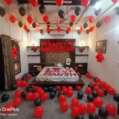 A personalized anniversary decoration in a bold red and black theme. This setup includes 'Happy Anniversary' foil balloons, the couple's names spelled out on the bed with petals, and balloons covering the floor and ceiling.