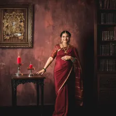 A solo portrait of a mother-to-be in a deep red saree, standing in a gracefully lit, vintage-themed studio setting that resembles a classic library.