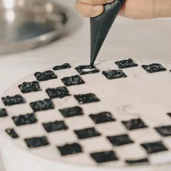 A chef carefully piping a checkerboard pattern with black squid ink. This shot captures the precision and artistic skill that goes into creating our visually striking dishes.