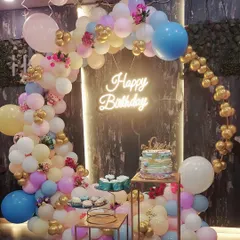 A colorful birthday party setup with a pastel-colored organic balloon arch and a bright 'Happy Birthday' neon sign. The design includes multi-level stands for the cake and desserts.