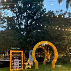 The rockstar party entrance at dusk, with string lights overhead creating a magical ambiance. The glowing marquee signs and arches stand out beautifully against the evening sky.