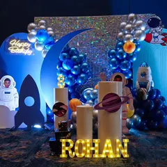 A cosmic adventure for Rohan's birthday with this outer space theme. The setup features a shimmering sequin backdrop, rocket ships, planets, and an astronaut cutout, all in a cool blue and silver color scheme.