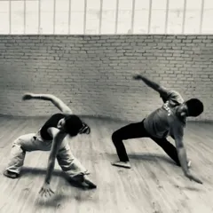 Two dancers in a powerful, grounded pose during a rehearsal for 'Dream Baby Dream'. The black and white filter highlights the raw physicality of the performance.