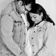 A casual and tender black and white photo. The couple, wearing matching denim jackets, shares a quiet moment, their heads together, focusing on the baby bump.