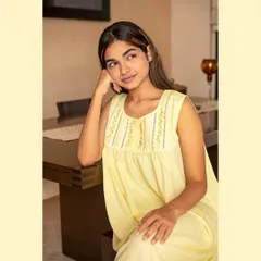 This image showcases a yellow cotton nightgown in a natural, daytime setting. The soft lighting and thoughtful pose highlight the delicate embroidery on the yoke, appealing to customers looking for comfortable, handcrafted sleepwear.