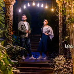 A cool, editorial-style portrait at night. The couple poses on the steps under hanging lights, framed by lush greenery, giving the shot a mysterious and cinematic vibe.