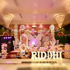 A grand first birthday stage for a butterfly and fairy theme. The setup includes large illuminated butterfly wings, marquee letters spelling "RIDDHI", and a beautiful pastel balloon arrangement, creating a stunning focal point in the banquet hall.