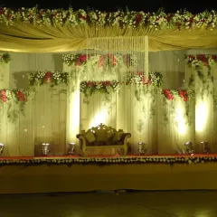 A classic stage decoration featuring a central crystal chandelier hanging from a draped ceiling. The backdrop is layered with fresh flowers in red and white, complemented by warm uplighting to create a grand and inviting focal point.