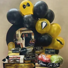 I created this luxurious "Jamai Raja" hamper to give the groom-to-be the royal treatment. It's packed with a mix of gourmet food platters like lasagna, fresh fruits, and sweets, all personalized with a photo and themed balloons in a sophisticated black and gold palette.