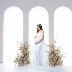 A serene and classic portrait of a mother-to-be in a sheer white gown. She stands between two floral arrangements against a clean, arched backdrop, creating an angelic look.