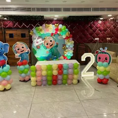 A colorful Cocomelon theme setup for a second birthday. This includes multiple character cutouts, a pastel balloon-wrapped table, and a light-up number '2'.