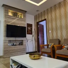 This living room showcases a custom TV unit clad in travertine stone, featuring built-in niches for electronics and decor. The design is complemented by a patterned wallpaper accent wall and a tray ceiling with recessed lighting, creating a warm and textured environment.