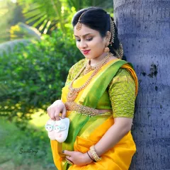 A beautiful outdoor portrait in a traditional yellow saree. The mother-to-be holds a pair of tiny shoes, standing by a palm tree in the warm, natural light.