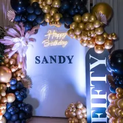 A chic and glamorous 50th birthday celebration setup. The black and gold balloon arch, custom 'Happy Birthday Sandy' backdrop, and 'FIFTY' signage create a sophisticated photo opportunity for a milestone event.