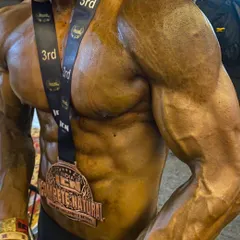 A close-up of a client's physique on show day, wearing his 3rd place medal. The vascularity and grainy muscle detail are signs of peak conditioning.