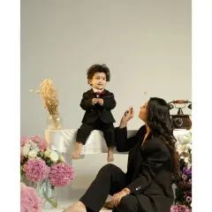 A playful and stylish studio shot. The little boy, dressed in a tiny suit, sits on a table while his mother engages with him from below. The scene is filled with charm, flowers, and a sense of fun.