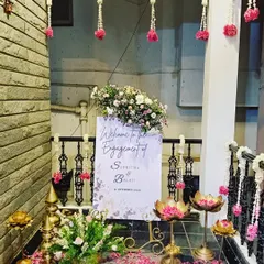 A charming welcome sign for an engagement ceremony. The sign is surrounded by traditional lotus-shaped brass lamps and hanging floral strings, creating a warm and inviting entrance for the guests.
