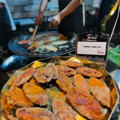 A close-up of our Surmai Tawa Fry, perfectly seasoned and pan-fried until golden brown. In the background, you can see the chef cooking more fresh fish on a large tawa.