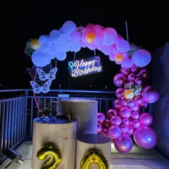 A 20th birthday celebration on a rooftop terrace at night. The setup features a balloon arch with a "Happy Birthday" neon sign, illuminated butterfly accents, and foil number balloons.