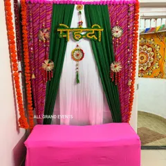 A classic Mehendi ceremony backdrop for home functions. We use a combination of green and pink bandhani-style drapes, bordered with marigold garlands, and a 'मेहन्दी' sign to create a festive photo corner.