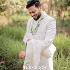A handsome portrait of the groom in his traditional white sherwani, adjusting his attire in a lush, green outdoor setting.