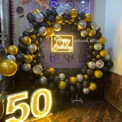 A stylish 50th birthday celebration with a black, gold, and silver theme. The decor features a circular balloon arch, a 'Happy Birthday' neon sign, and large marquee numbers.