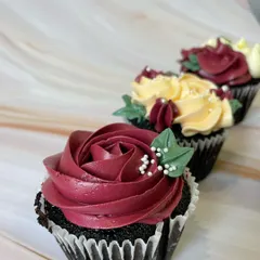 A close-up of a chocolate cupcake with a beautifully piped burgundy buttercream rose. The small green leaves and white sprinkles add a realistic and delicate touch to this mini dessert.