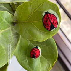 A top-down view of the hand-stitched ladybug badges on a vibrant green leaf. The contrast between the red thread and the natural background really makes the embroidery pop. These are such fun, whimsical pins.